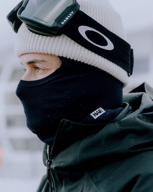 Person trägt HAE The Stormerer Sturmmaske in Schwarz mit Mütze und Skibrille im Schnee – winddichte Balaclava für Skitouren